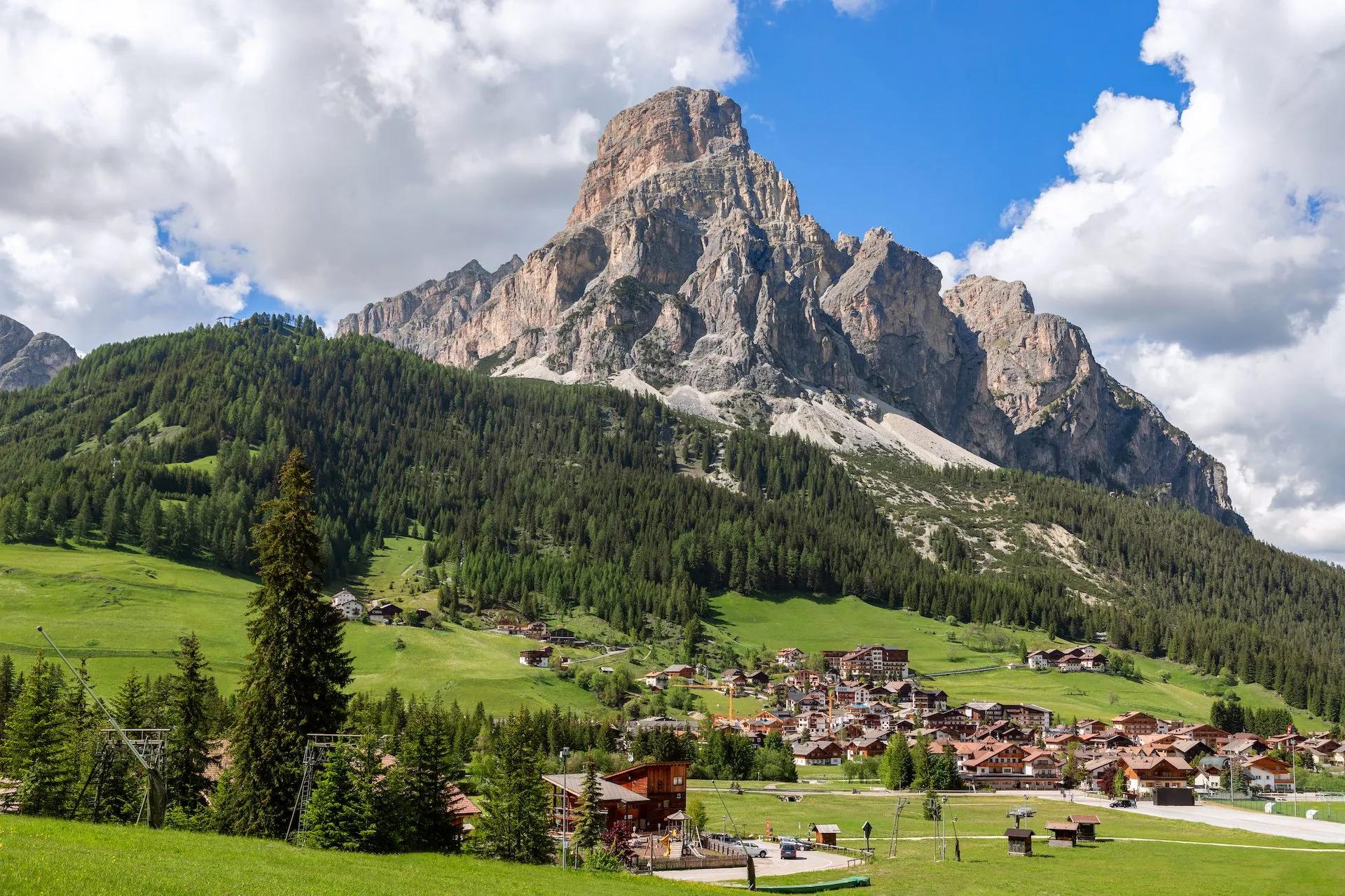 Alpine village nestled in green valley below massive rocky mountain under blue sky with clouds.
