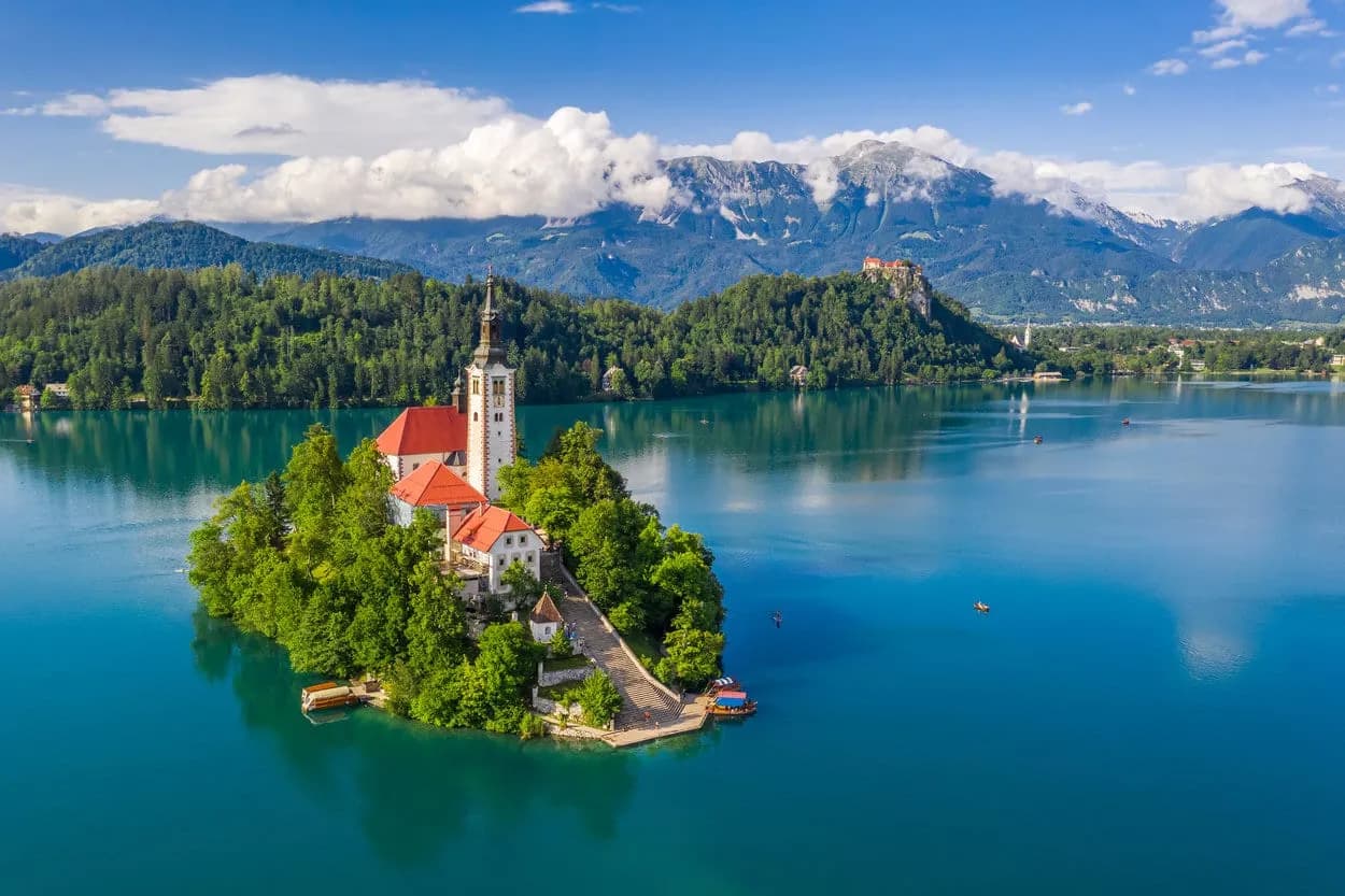 Church on island in Lake Bled with Julian Alps and castle on forested hill.