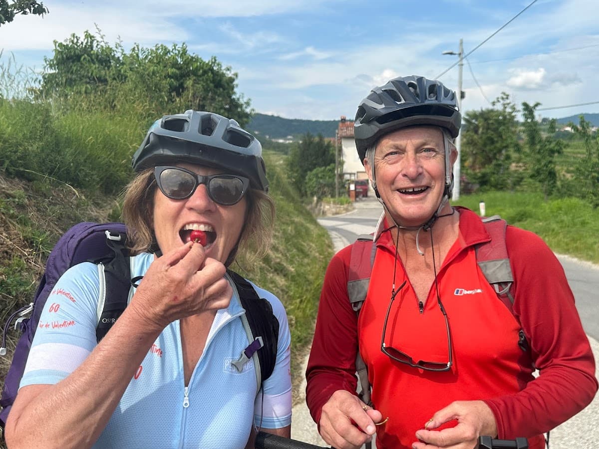Two cyclists wearing helmets take a break on a rural road, one eating a cherry.