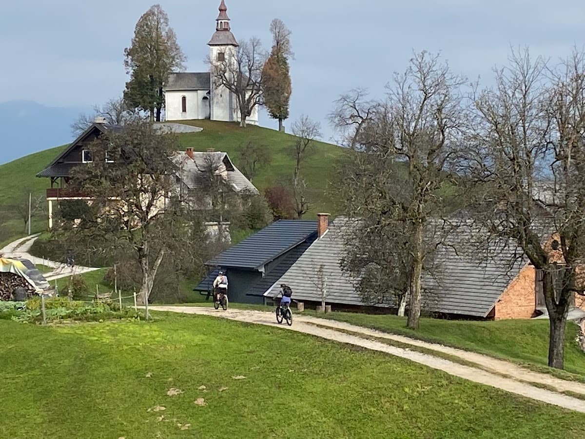 Two cyclists ride on a dirt path past houses toward a white church on a grassy hill.