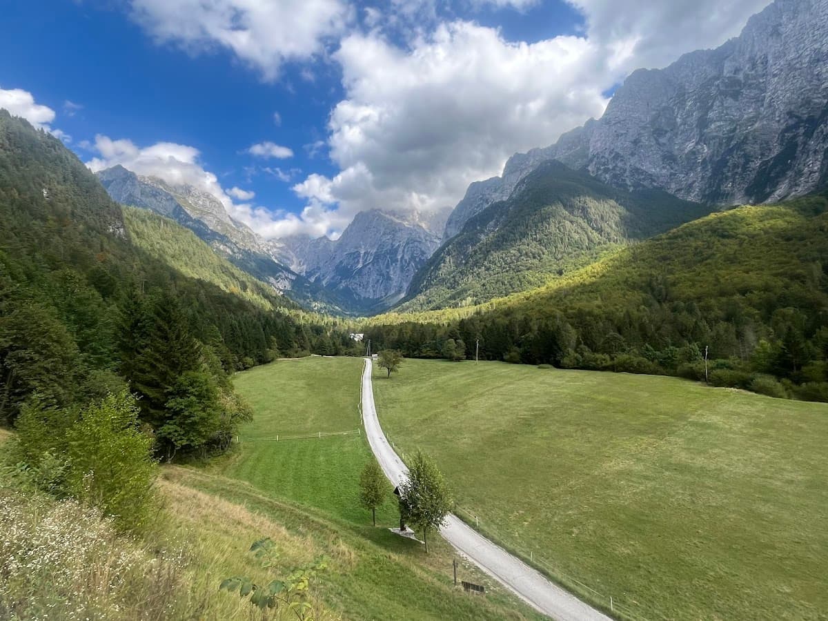 Winding road through green valley floor surrounded by steep, forested mountains under a partly cloudy sky.