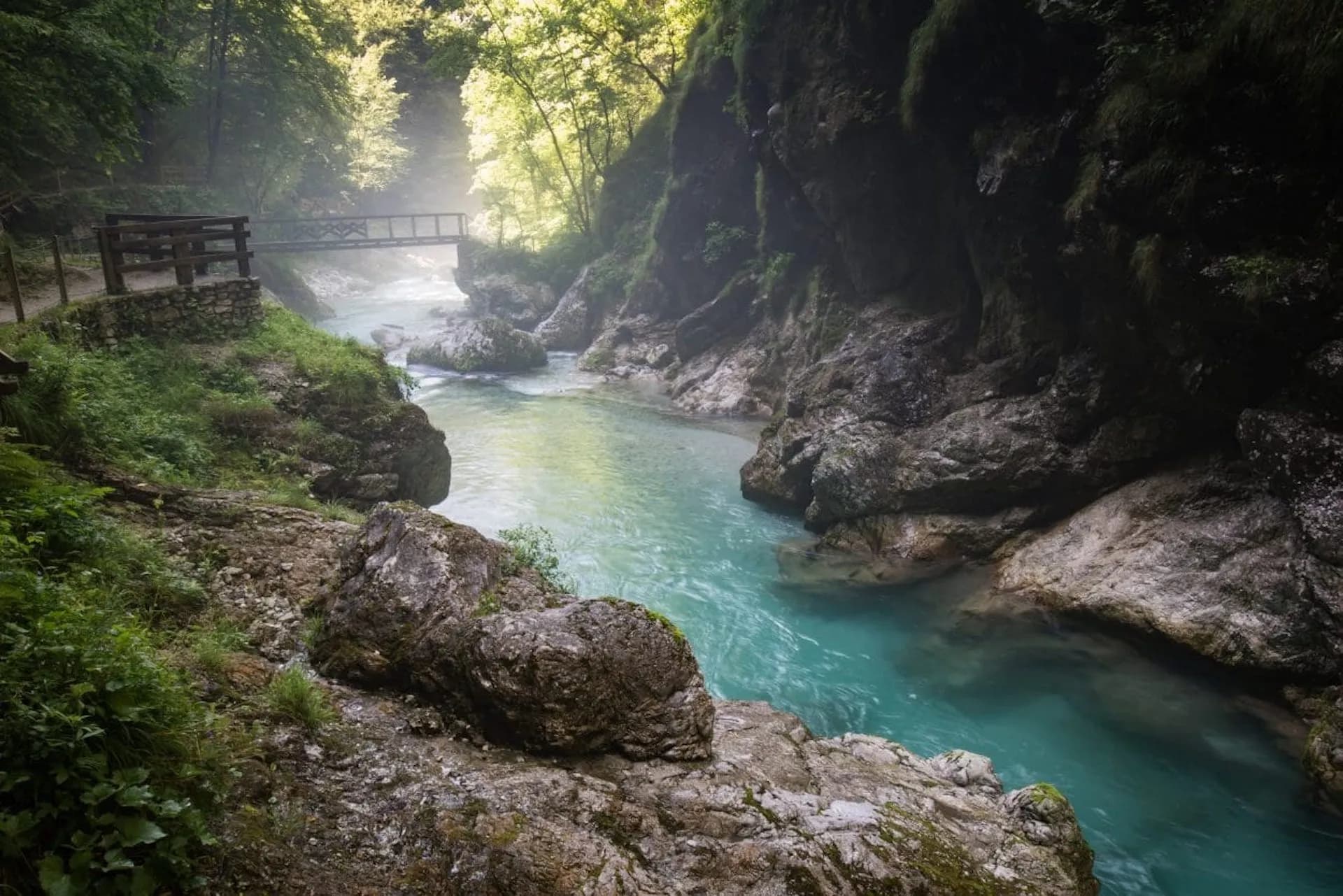 Turquoise river flowing through a rocky gorge with a wooden footbridge and lush green forest.