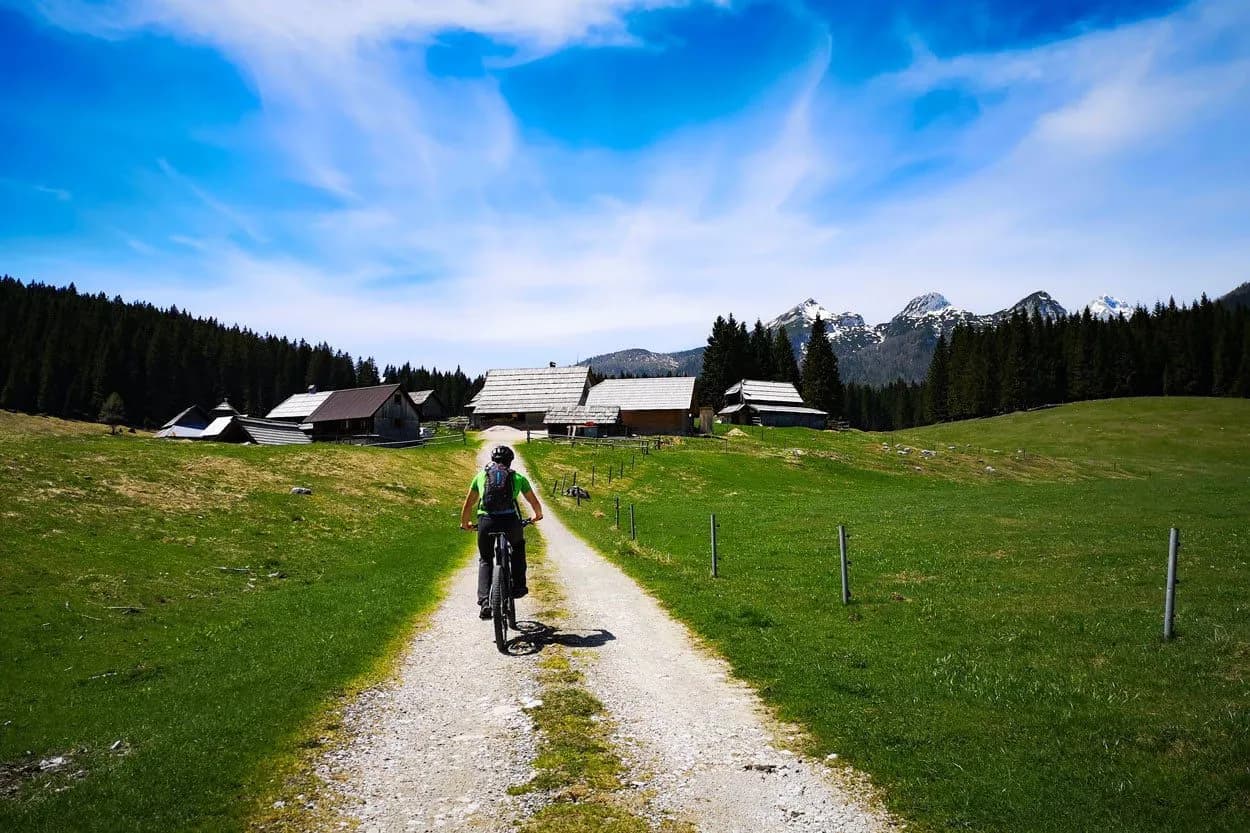Mountain biking on gravel path toward wooden huts near snowy peaks in Pokljuka Zajavornik.