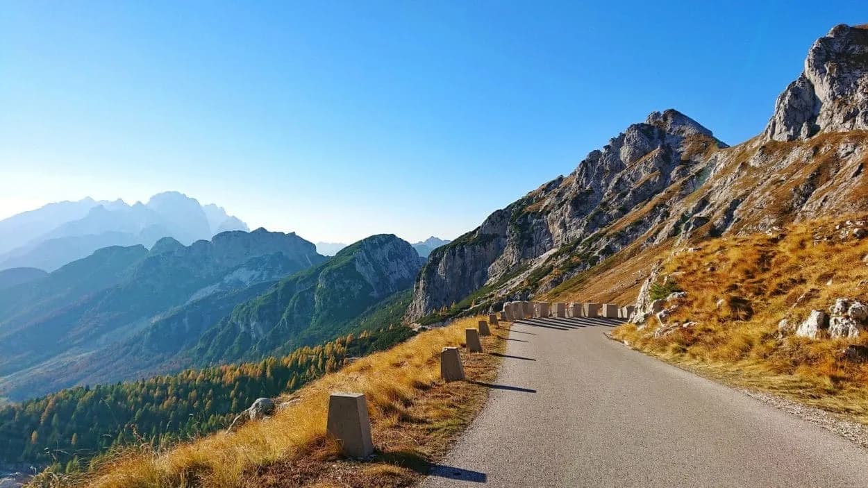 Winding mountain road ascending past rocky slopes with autumn grass and distant peaks.