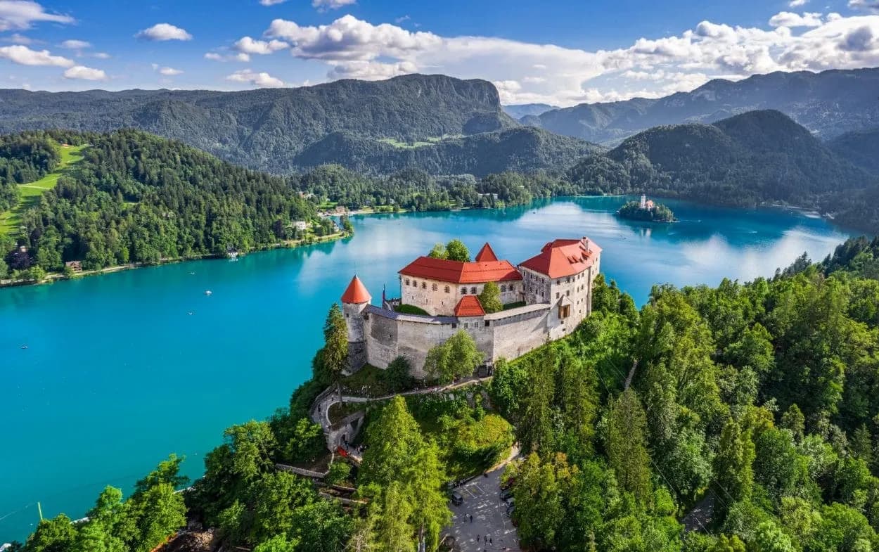 Lake Bled Castle on cliff overlooking bright blue lake with forested mountains and island church.