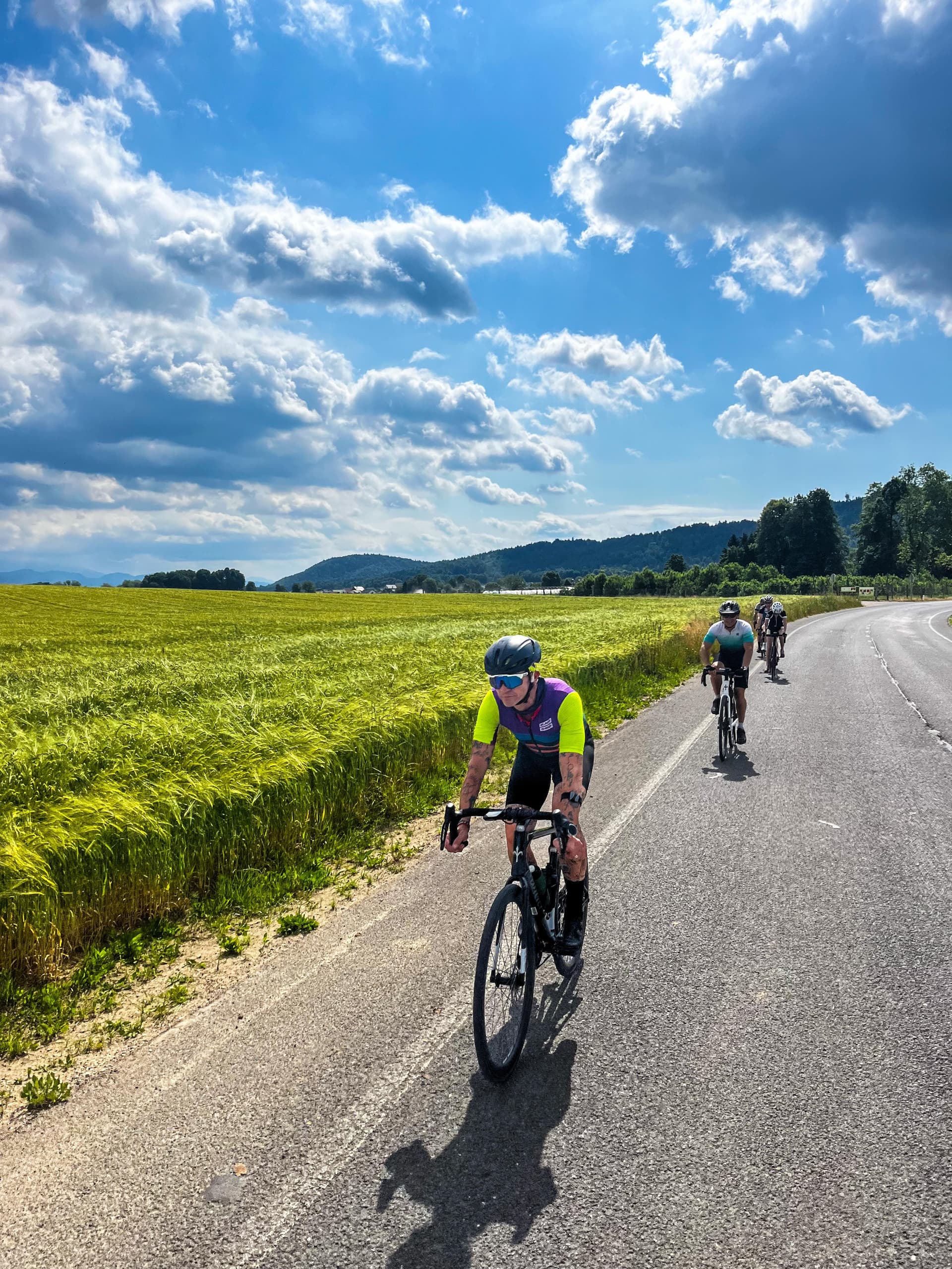 Cyclists riding on road beside bright green field under dramatic blue sky in Slovenia.