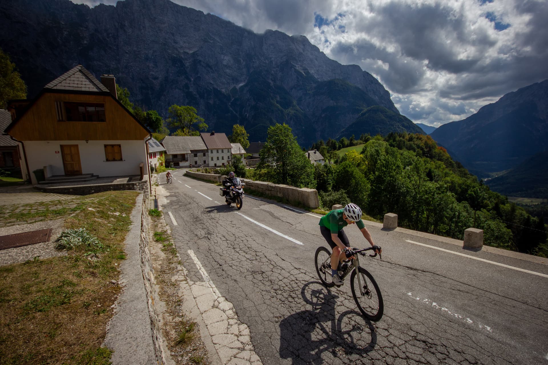 Cyclist riding uphill past houses with a motorcycle and massive mountains under cloudy sky.