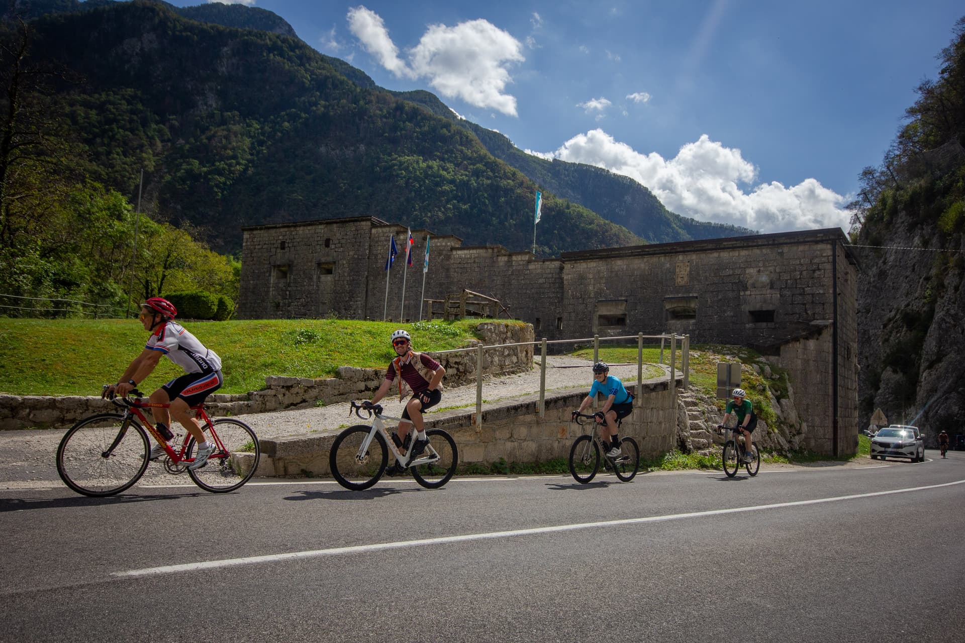 Cyclists riding past Fort Kluze in Slovenia with steep, forested mountains.