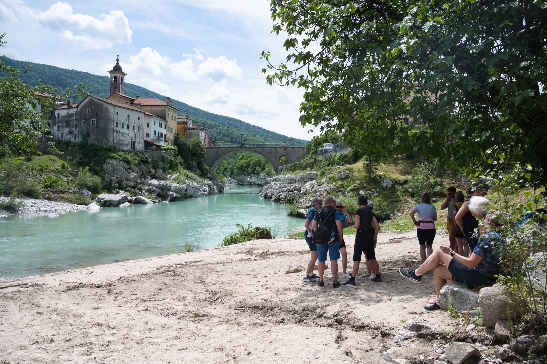 Hikers resting on sandy bank of turquoise river near stone bridge and town in Solkan.