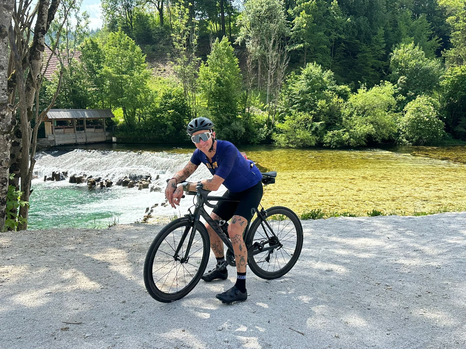 Cyclist with road bike resting by a river with a small wooden structure and lush green trees.