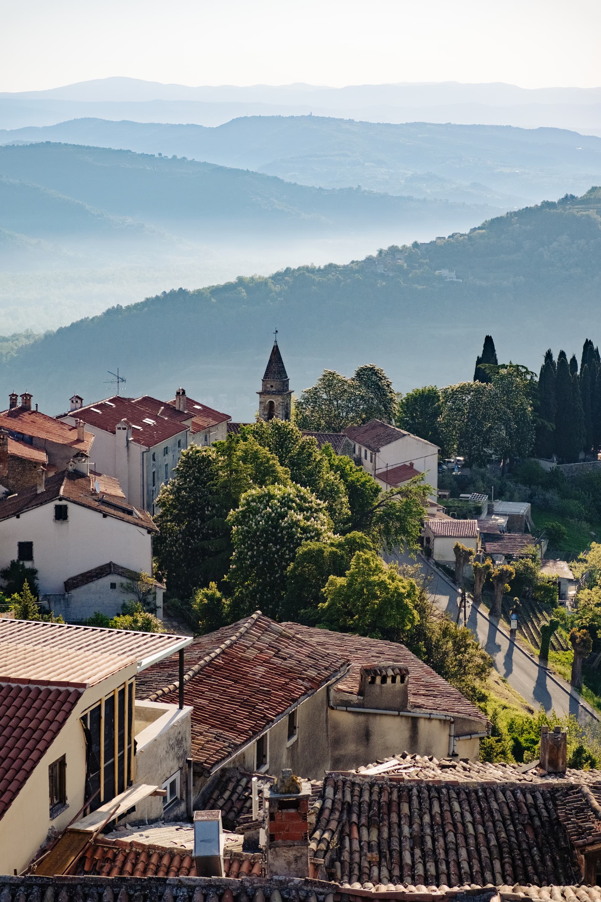 Hillside village with terracotta roofs and church tower overlooking hazy blue mountains.