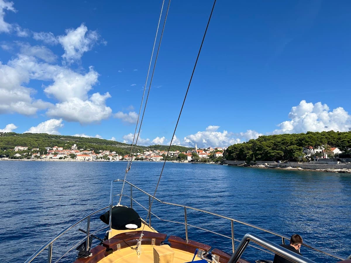 View from sailboat bow approaching coastal town with white buildings and green hills under blue sky.
