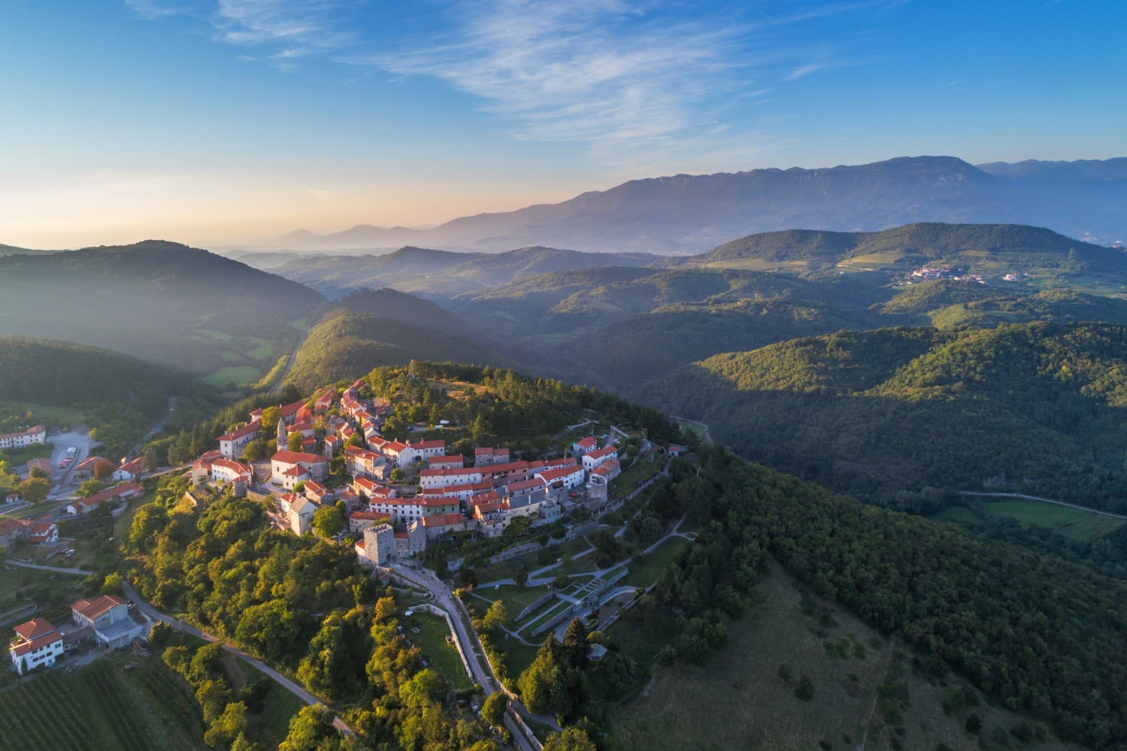 Aerial view of Štanjel hilltop village with stone houses and red roofs surrounded by green hills at sunset.