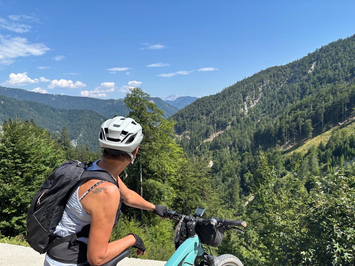 Cyclist wearing a helmet and backpack looking out over a mountain valley with forest and blue sky