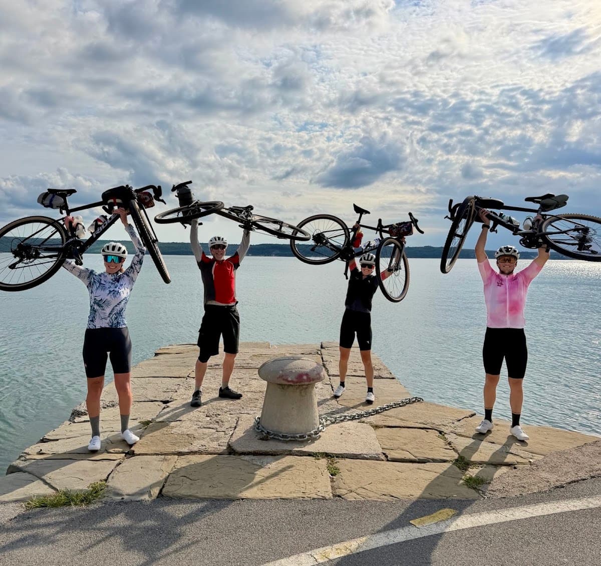 Four cyclists holding bicycles high above their heads on a stone pier by the water