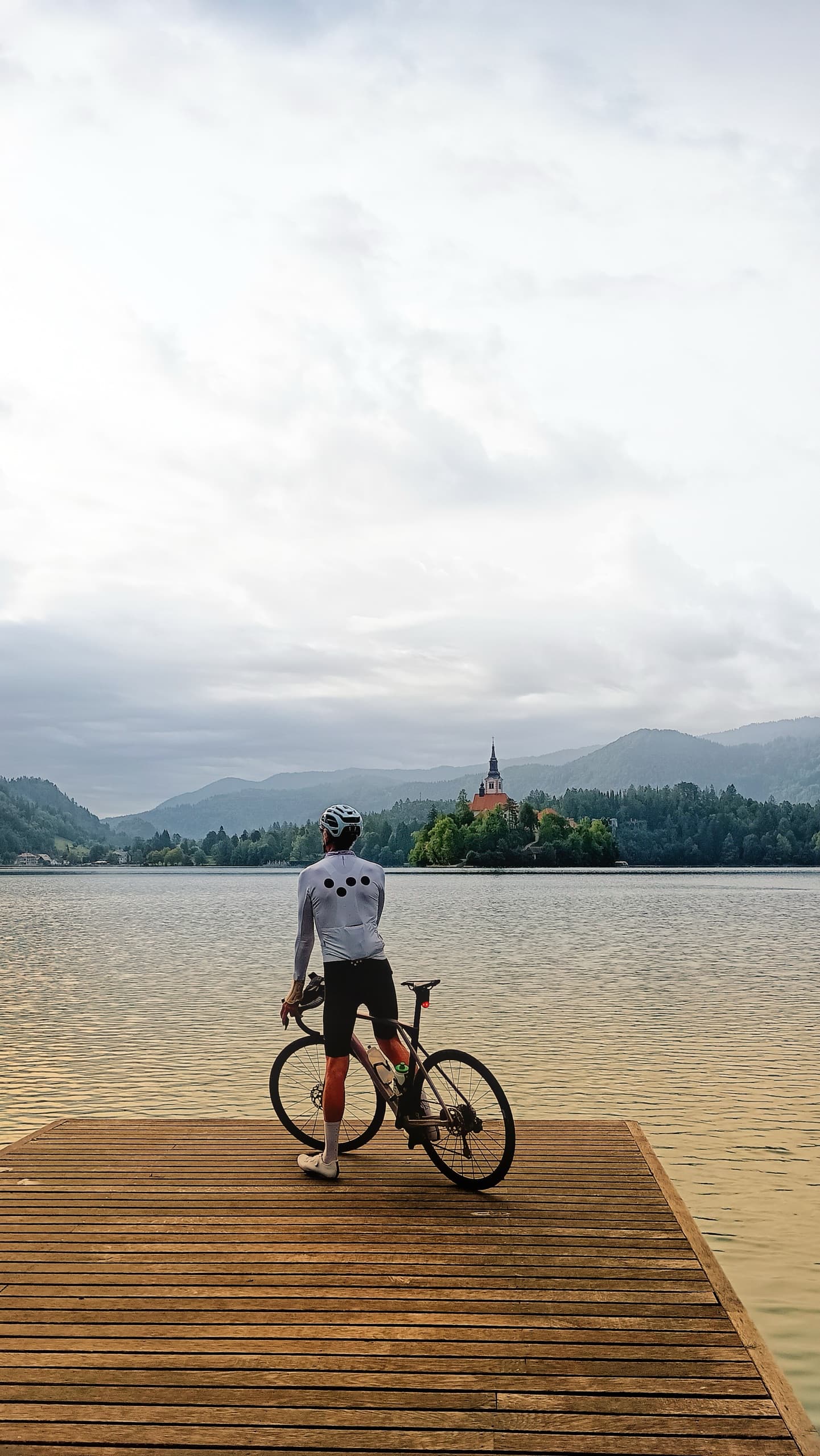 Cyclist standing on a wooden jetty at Lake Bled with island church and mountains