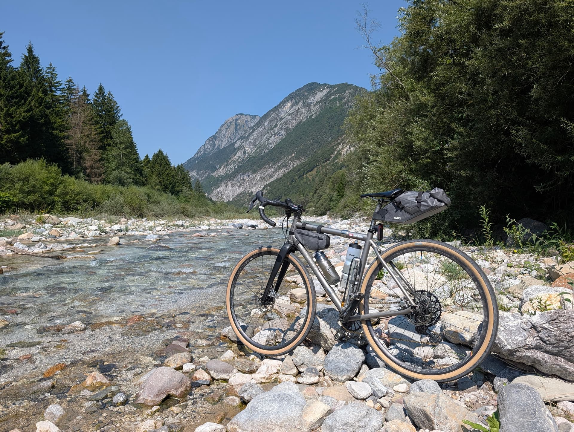 Gravel bike parked on a stony riverbed below mountain peaks and pine trees