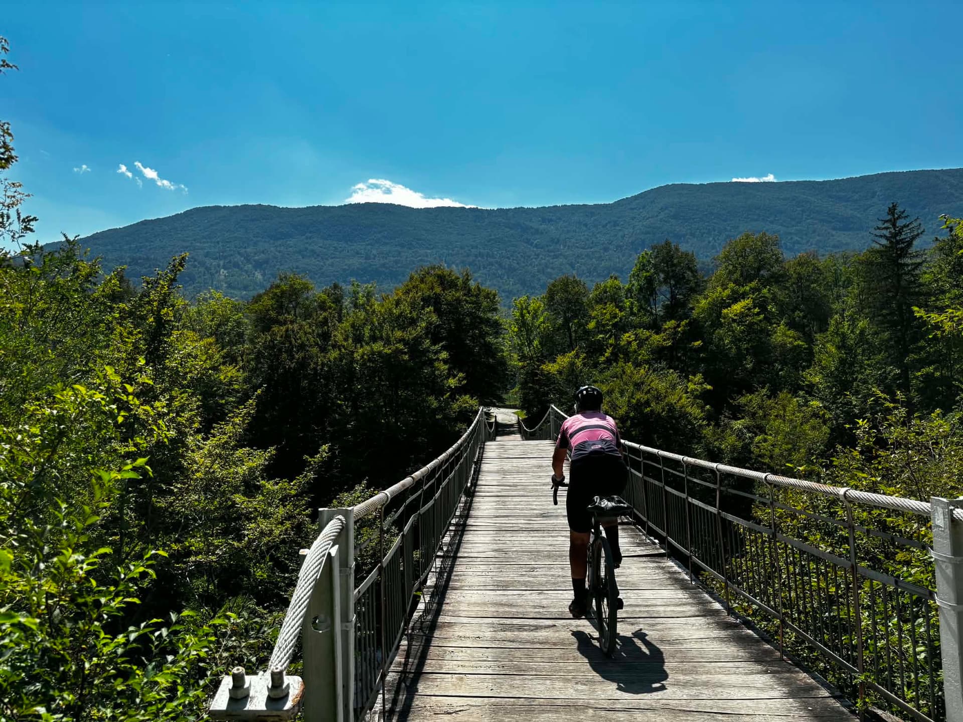 Cyclist riding across a wooden suspension bridge in a forest with mountains in the background