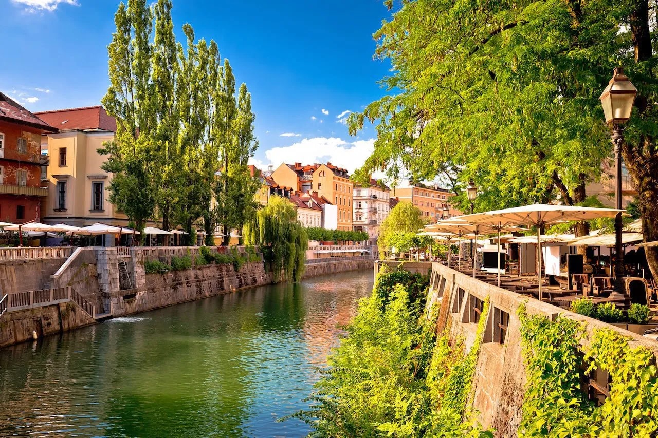 River lined with stone embankments, lush trees, and outdoor cafe seating in sunny Ljubljana.
