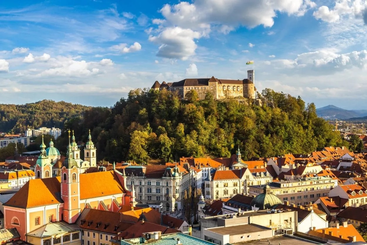 Ljubljana Castle atop a wooded hill overlooking city rooftops and a pink church.