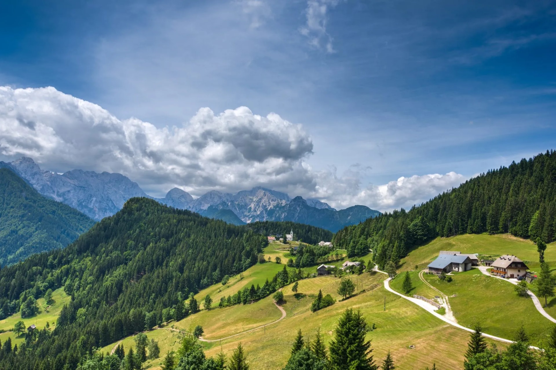 Panoramic summer road through green alpine meadows with distant rugged mountains under clouds