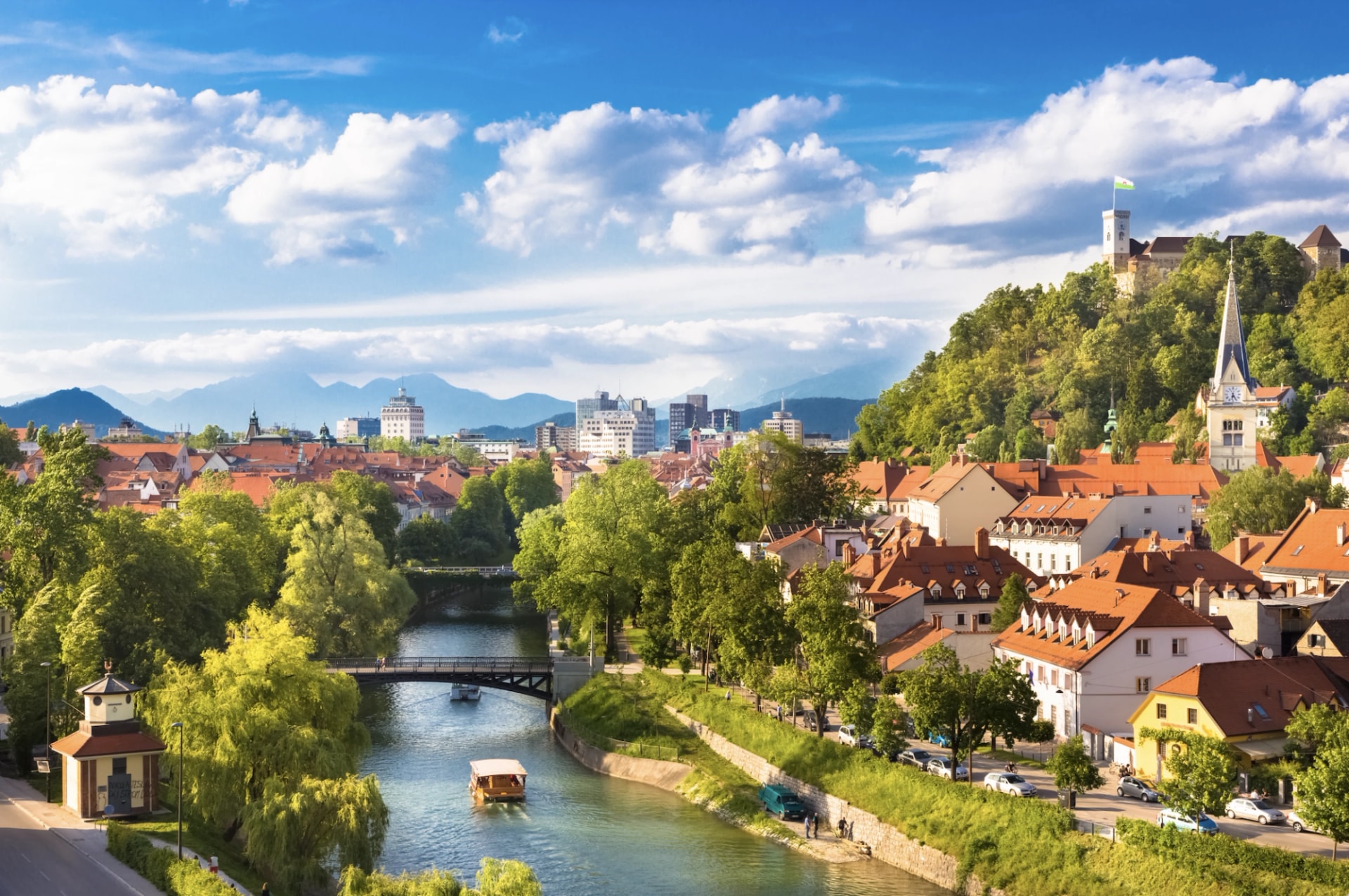Ljubljana cityscape with riverboats, castle on a hill, and Julian Alps in the background.