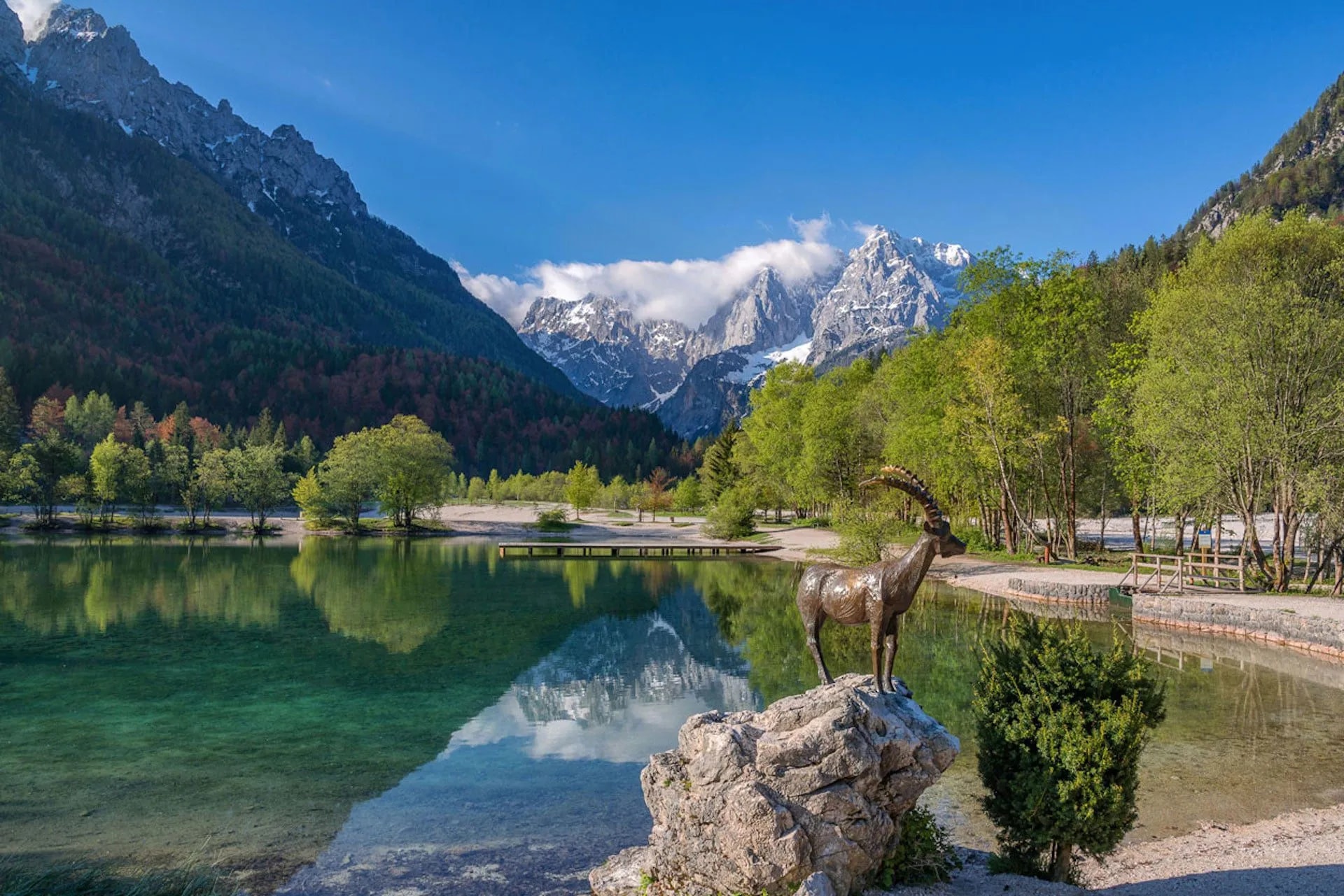 Ibex statue by clear alpine lake reflecting snow-capped mountains and green forest