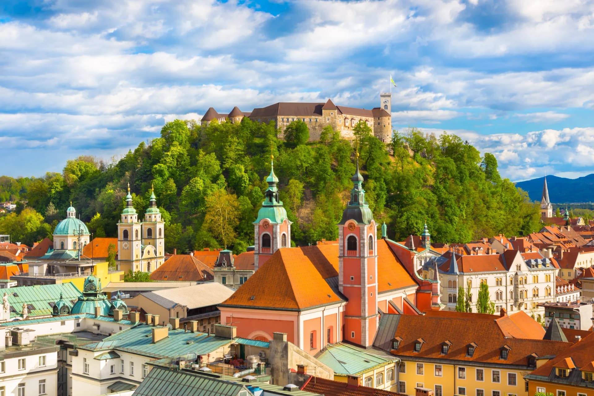 Rooftops of Ljubljana with a castle on a green hill under a blue cloudy sky.