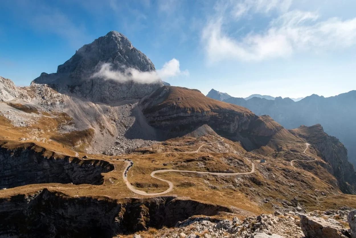 Winding mountain road ascending past rocky slopes toward a jagged peak under a blue sky.