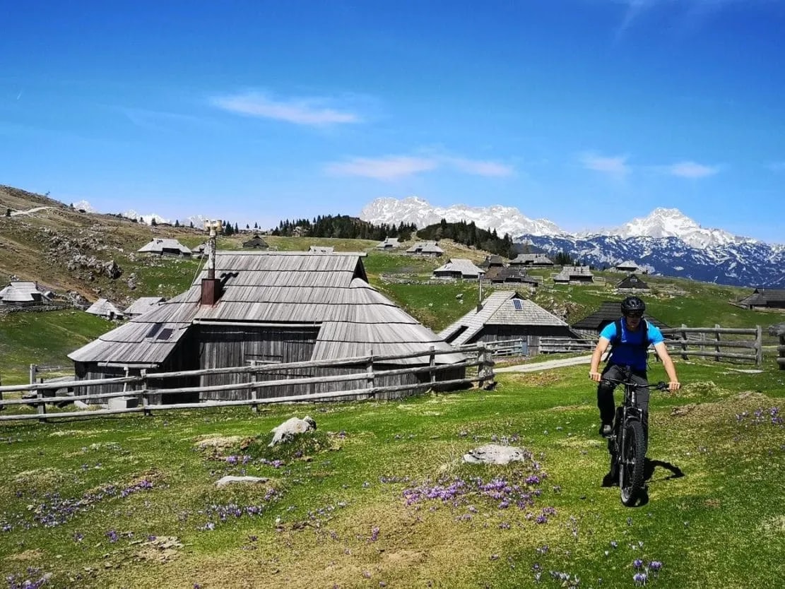 Mountain biking past wooden huts on Velika Planina with snow-capped mountains in background.