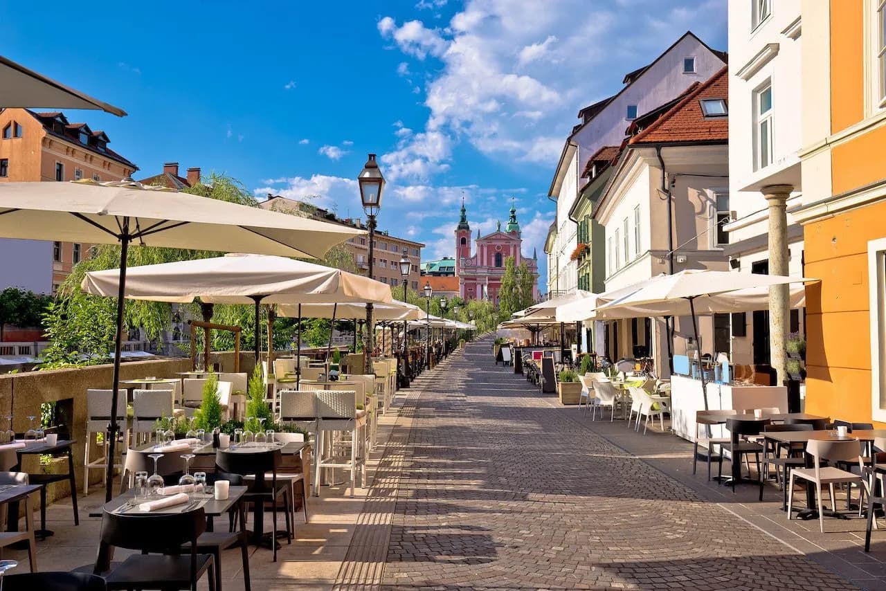 Outdoor cafe seating on cobblestone street toward pink church in Ljubljana Center.