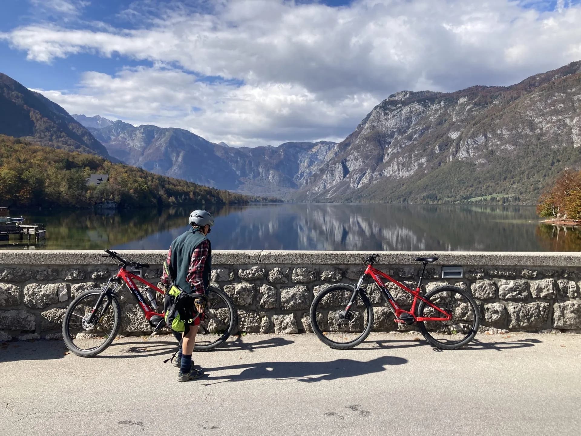 Cyclist with two mountain bikes by stone wall overlooking Lake Bohinj and alpine mountains.