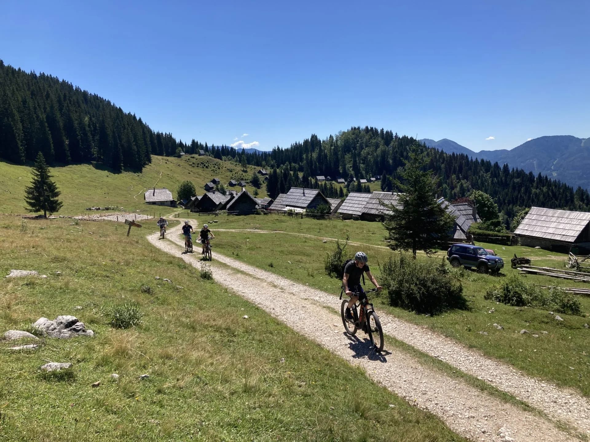 Cycling along a gravel path past traditional wooden houses in a sunny mountain pasture.