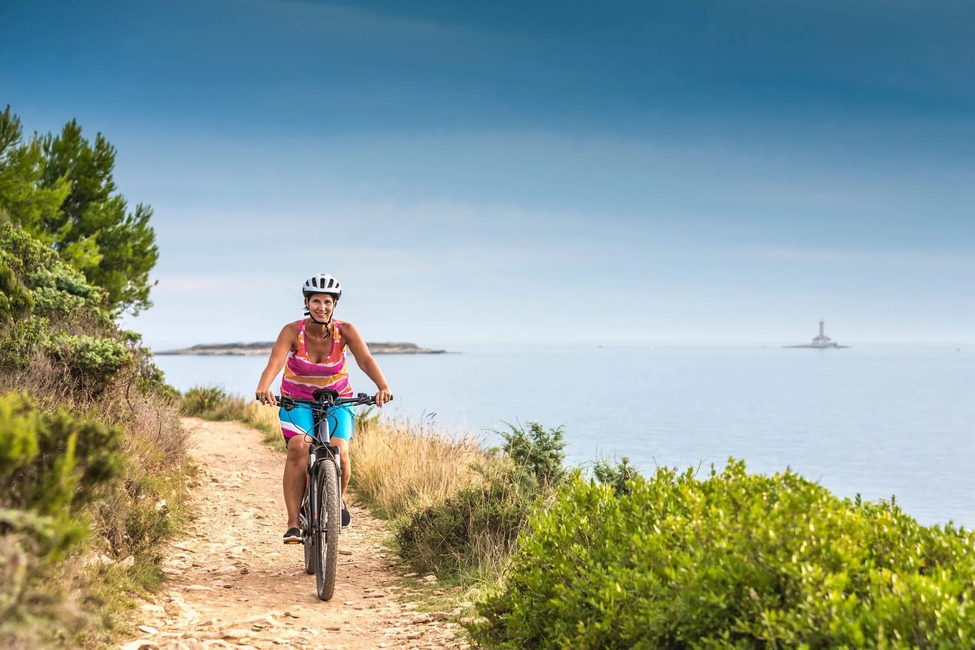 Woman cycling on dirt path along the Kamenjak Cape coastline with a lighthouse offshore.