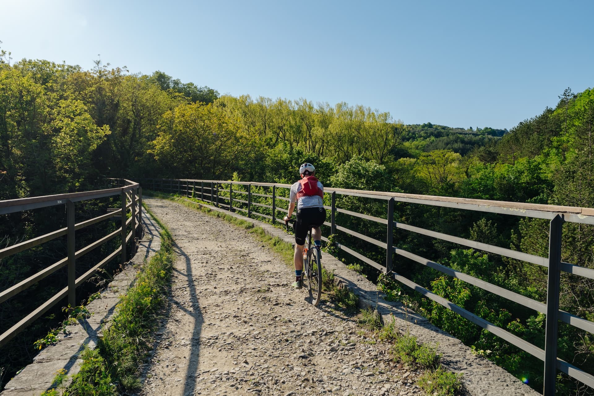 Cyclist with red backpack riding on Parenzana trail bridge over lush green valley.