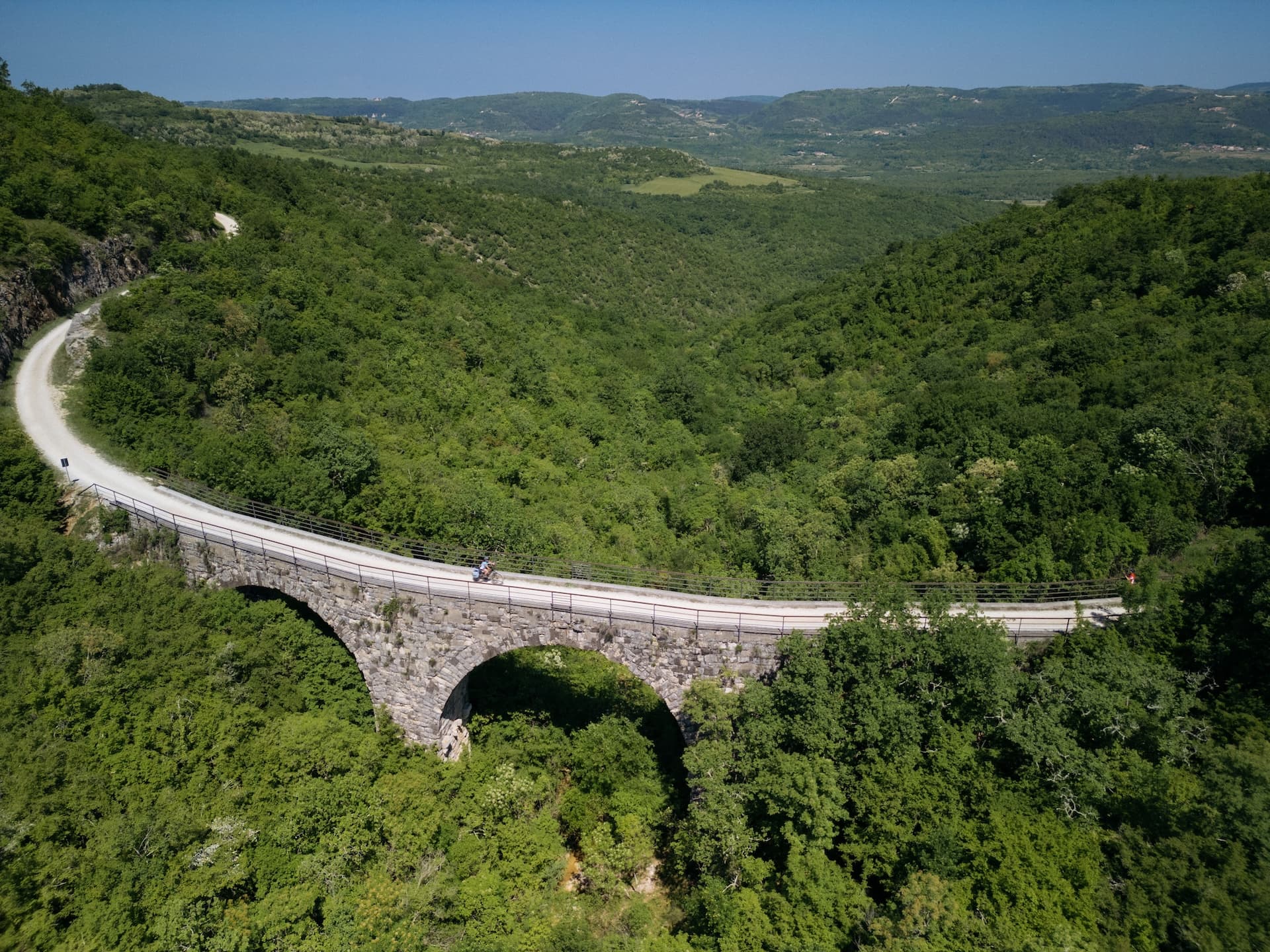 Cyclist crossing stone Parenzana bridge over dense green valley landscape