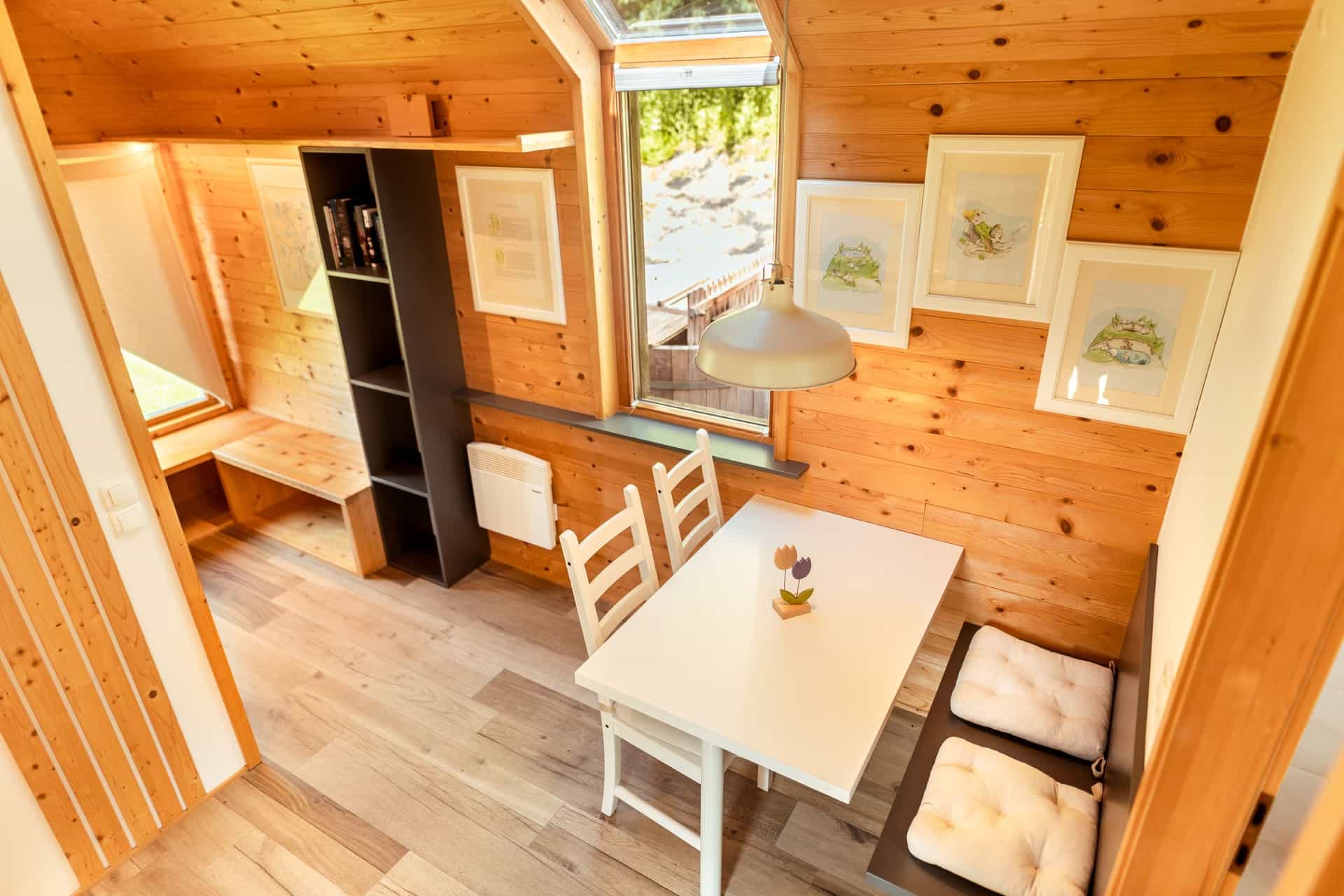 Cozy wooden interior with dining table, white chairs, and framed art near a bright window.