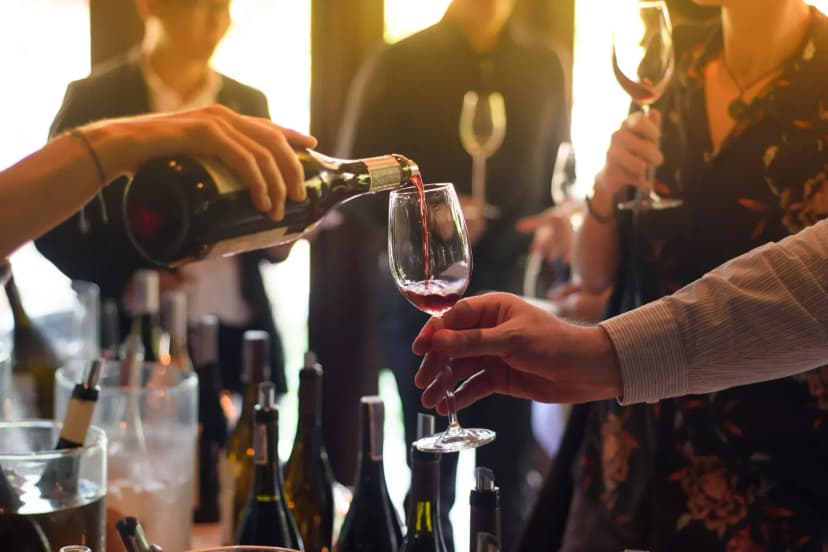Pouring red wine from a bottle into a glass during a tasting event with people in the background.