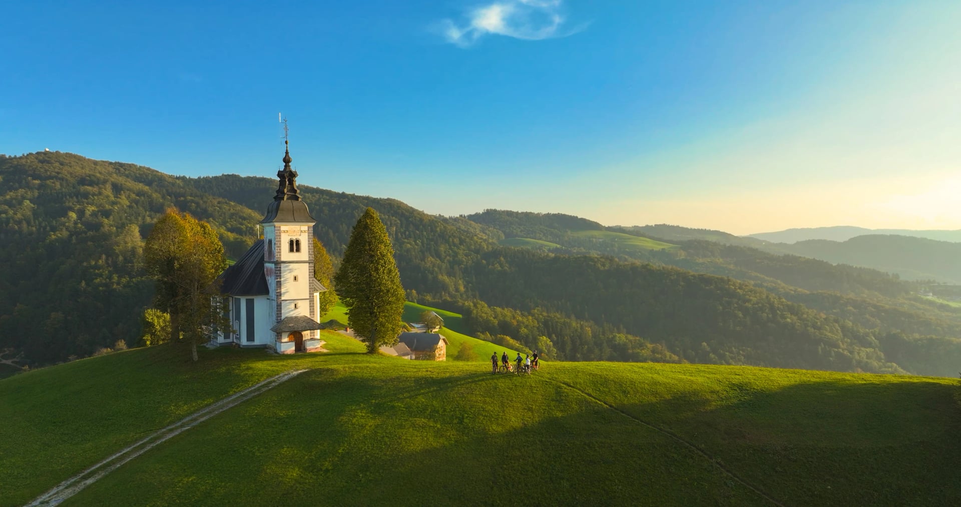 AERIAL: Group of mountain bikers by the scenic old church on top of the hill, surrounded by lush green mountains and bathed in late afternoon sunlight. Nice day for biking in the hilly countryside.