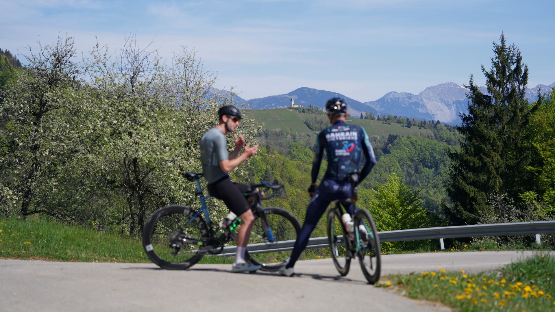 Cyclists resting on a road with spring blossoms and mountain views.
