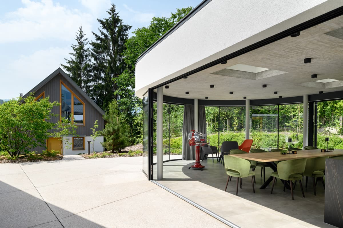 Dining interior in a modern glass pavilion with green chairs and a wooden table, set in a forest landscape.