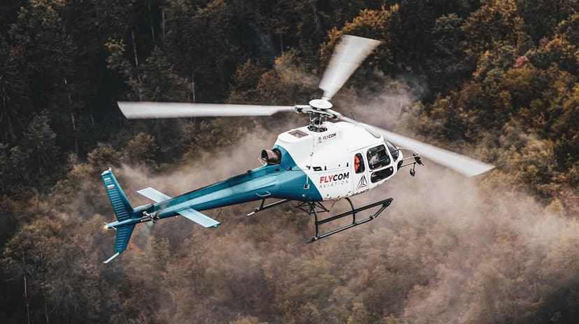Flycom helicopter flying over a forest with autumn foliage.