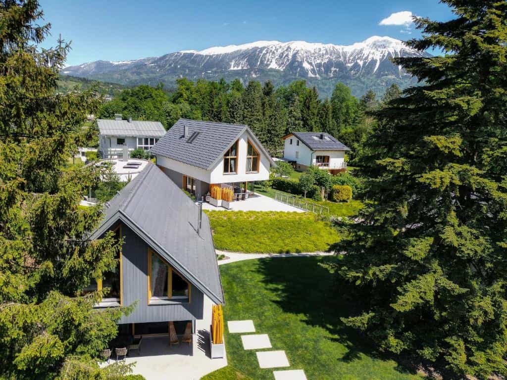 Modern resort villas with grey roofs in a lush green valley beneath snow-capped alpine mountains.