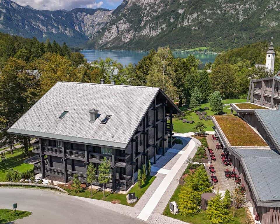 Modern wooden hotel building near a lake with a church steeple, surrounded by mountains and forest.
