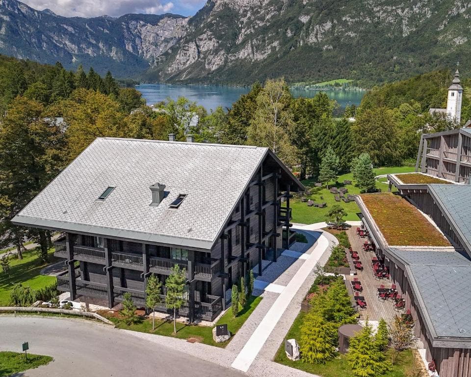 Modern wooden hotel building near a lake with a church steeple, surrounded by mountains and forest.