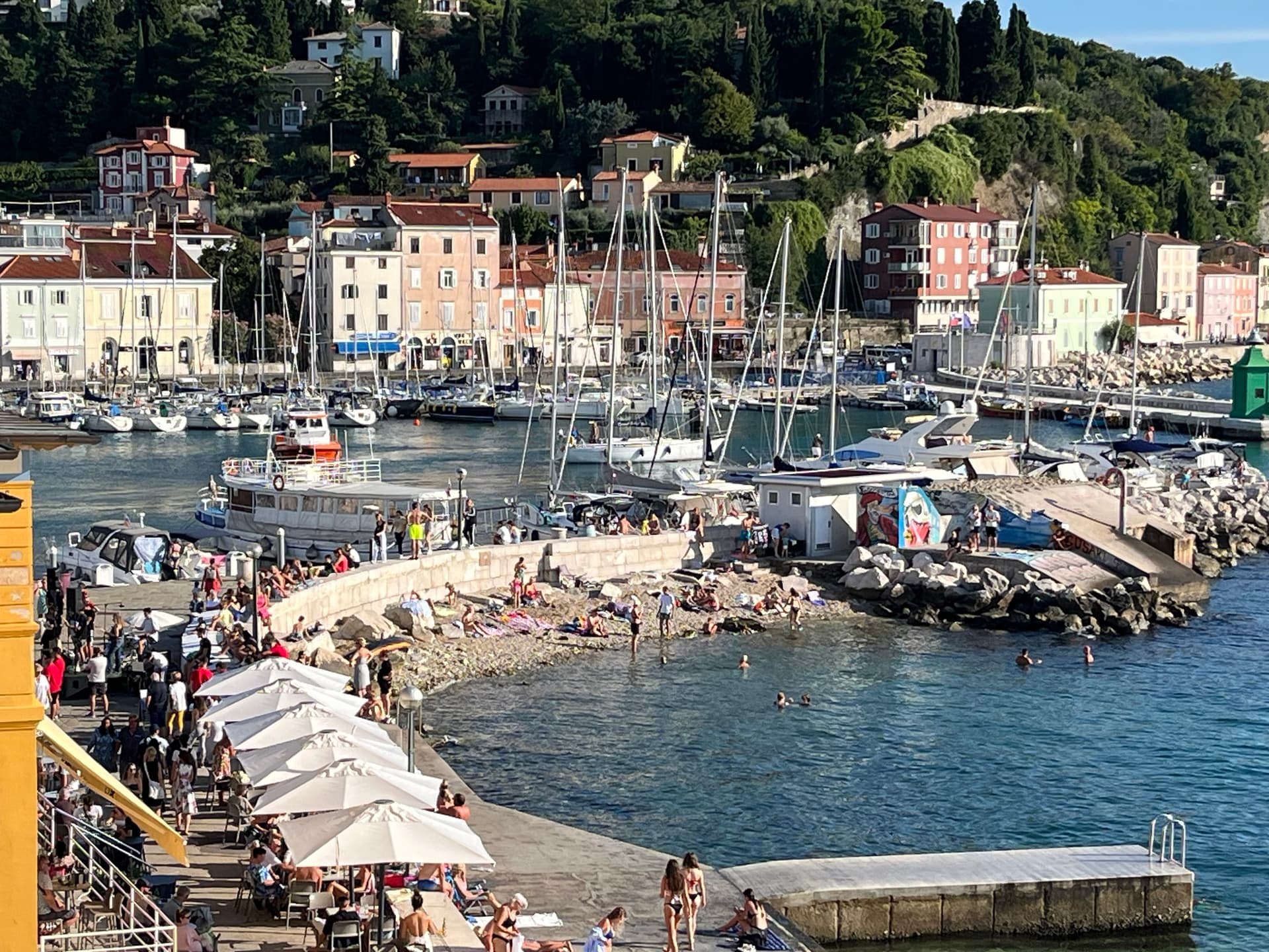 Piran harbor and seafront with moored boats, beachgoers, swimmers, and outdoor dining umbrellas